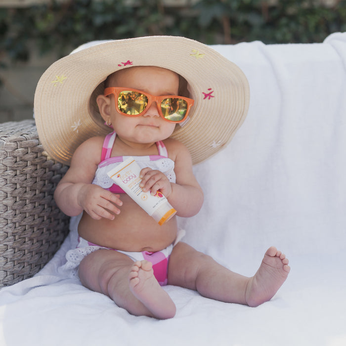 Baby in swimsuit, sunglasses and sun hat holding Non-Toxic, Hypoallergenic Broad Spectrum SPF 30 + Sunscreen