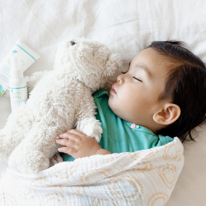 Sleeping baby swaddled with teddy bear and Non-Toxic, Hypoallergenic Aromatherapy Calming Mist on a bed